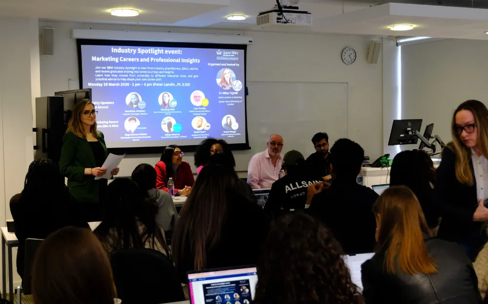 An image of students in a classroom looking at a screen with a powerpoint on it and a female speaker standing along with guest speakers sitting down