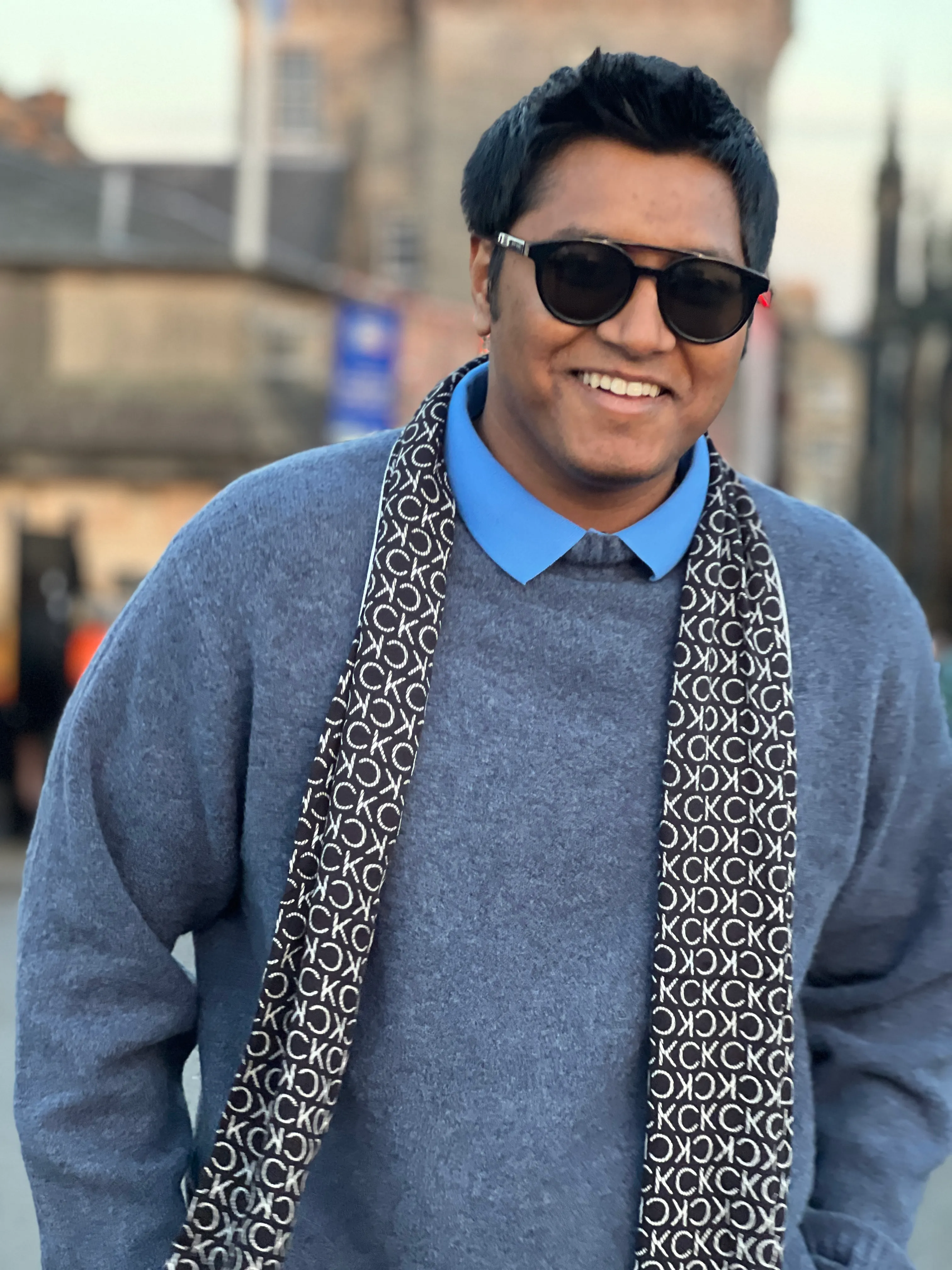 An image of a man wearing sunglasses and a scarf in a blue shirt and blue jumper outside smiling
