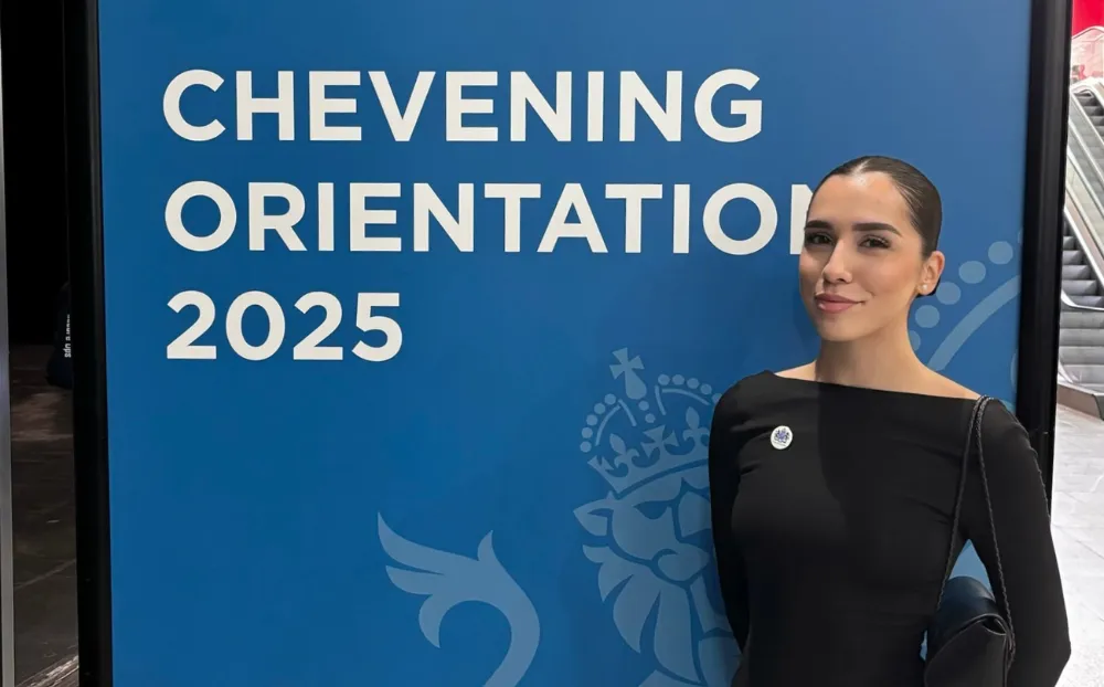 Image of a woman standing smiling wearing all black and a slick bun infront of a sign with text on it.