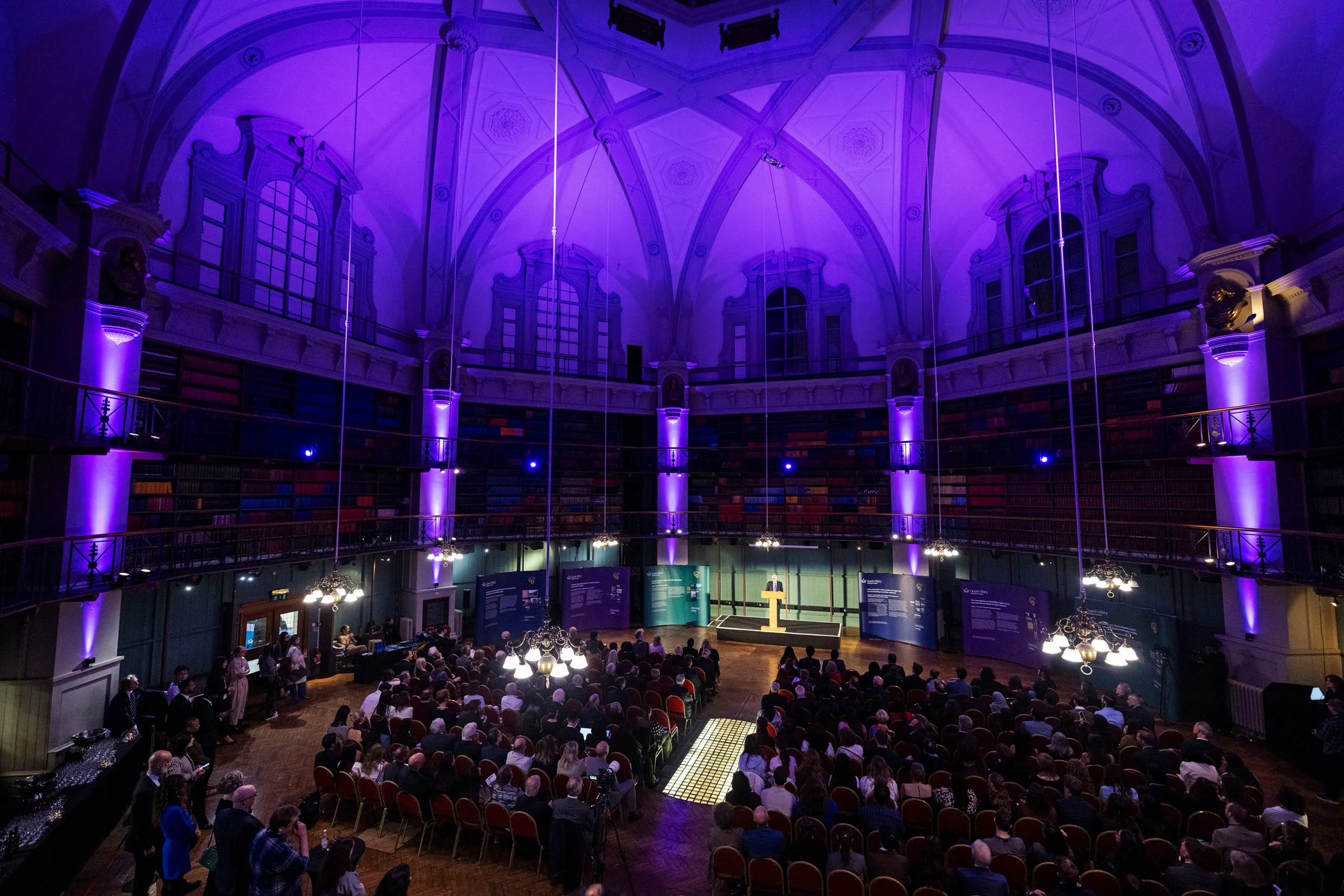 Octagon room lit in purple filled with an audience listening to a speaker on stage.