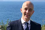 Ioannis Kokkoris wearing a suit and blue tie, standing in front of the ocean