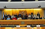 Malgosia Fitzmaurice and Andrew Pitts with Queen Mary LLM students on the stage of the the International Maritime Organization's assembly room. Behind them is the IMO logo on a wood paneled wall.