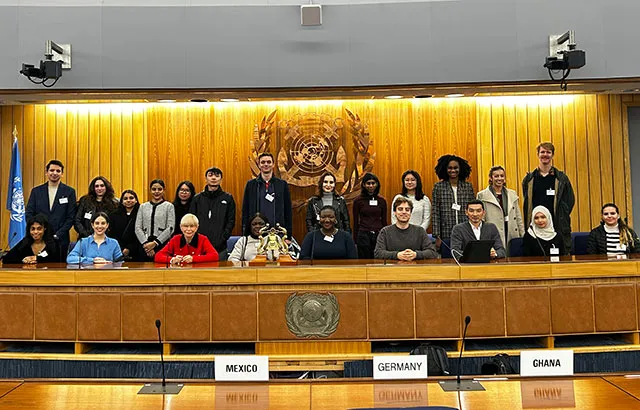 Malgosia Fitzmaurice and Andrew Pitts with Queen Mary LLM students on the stage of the the International Maritime Organization's assembly room. Behind them is the IMO logo on a wood paneled wall.