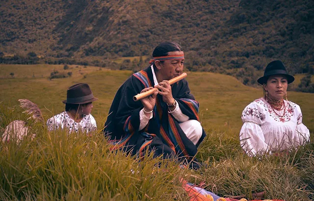 A man sat on the grass playing a wind instrument with a woman and child. They are wearing traditional dress.