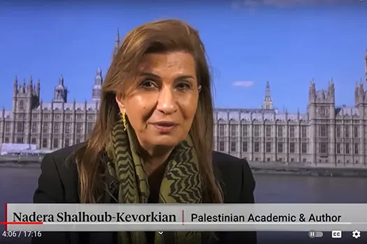 A still of Professor Nadera Shalhoub-Kevorkian speaking to Democracy Now! with the UK Houses of Parliament as a backdrop. She is wearing a black jacket, green scarf, and gold earrings.