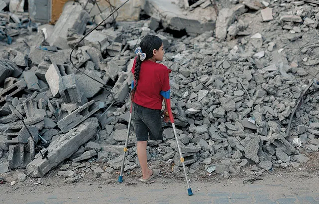 A Palestinian child standing in front of rubble in Gaza. She is missing her right leg and using crutches.