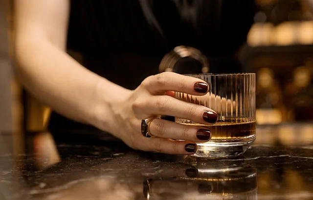 A person, presumably a woman, with their nails painted black and drinking a glass of whiskey in a bar.