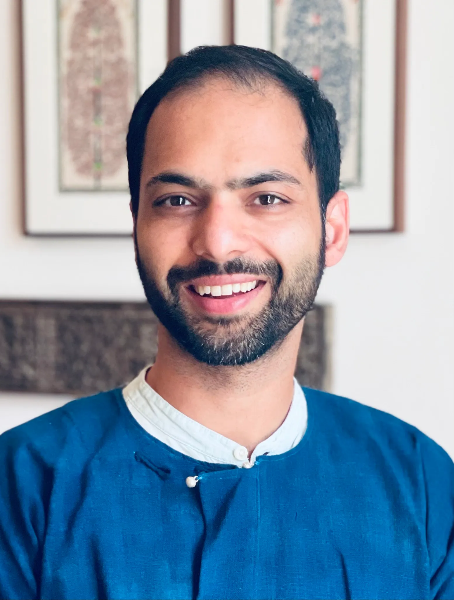 Mohsin has dark hair and a beard. He is smiling at the camera wearing a blue shirt