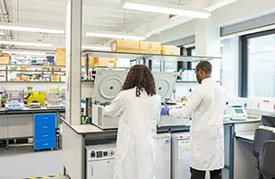 male and female student in lab coats stood by machinery in lab