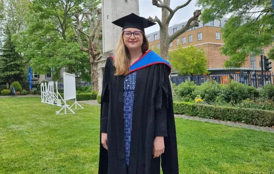 A woman posing for a photo in her graduand cap and gown outside the Queens Building of Queen Mary University.