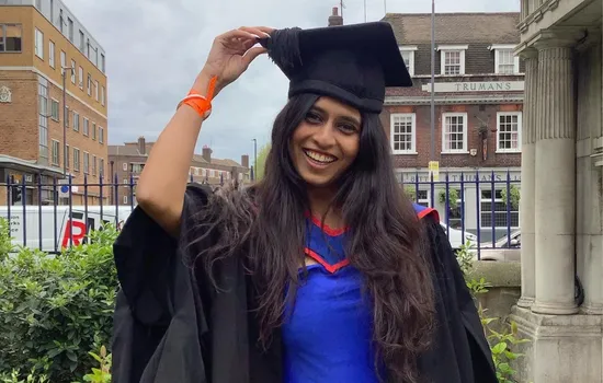 A young woman smiles and poses in her graduate cap and gown at the Mile end campus.