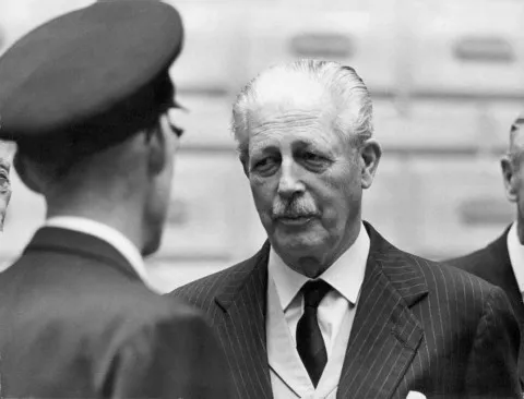 Conservative Prime Minister on a visit to Stockholm in 1963. Macmillan is wearing a dark suit and tie, with a pearl-coloured waist coat. He is looking at a military guard who has his back to the camera.
