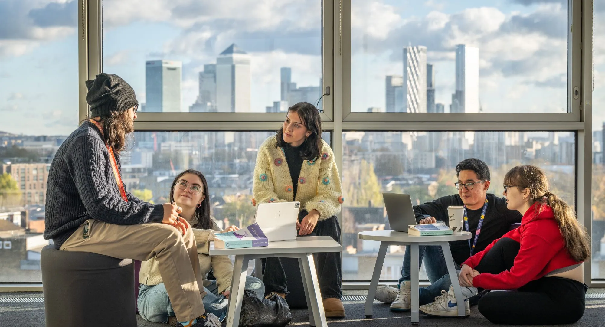 A group of students sit working in front of a window with skyscrapers in the city visible in the background