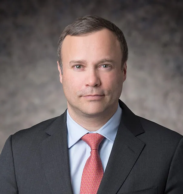 Dr Remy Gerbay in a grey suit, red tie and blue shirt against a grey bacground