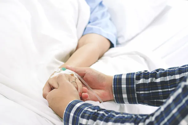 Close up of arm of patient in hospital bed, holding hands with someone next to the bed
