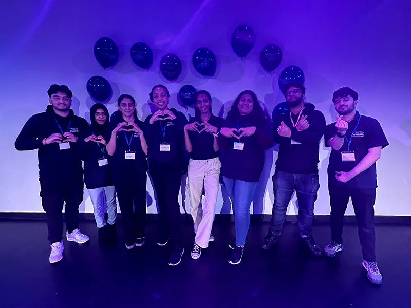 Student ambassadors making a heart sign with their hands, there are balloons behind them