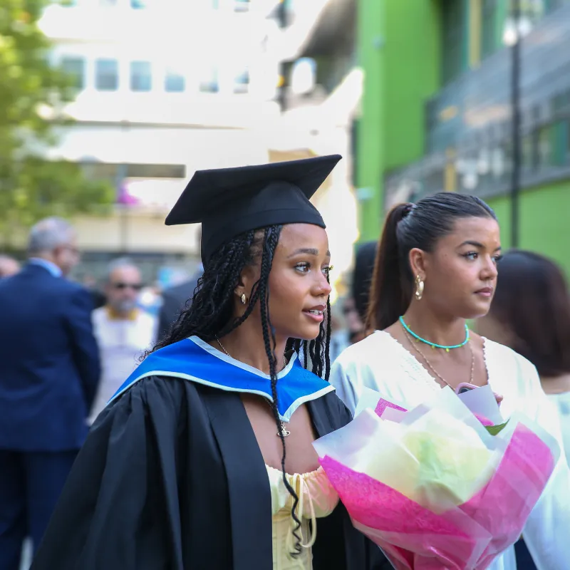 An image of a student wearing a graduation cap and gown and holding flowers looking away with another person behind her dressed formally also looking away.