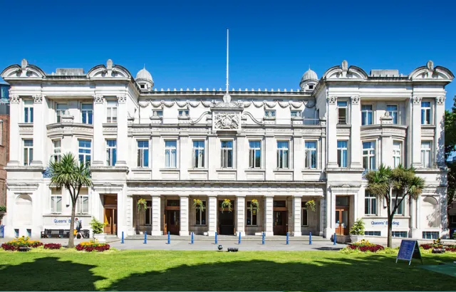 Picture of the neoclassical Queen's Building with a foreground of green grass and under a bright blue sky.