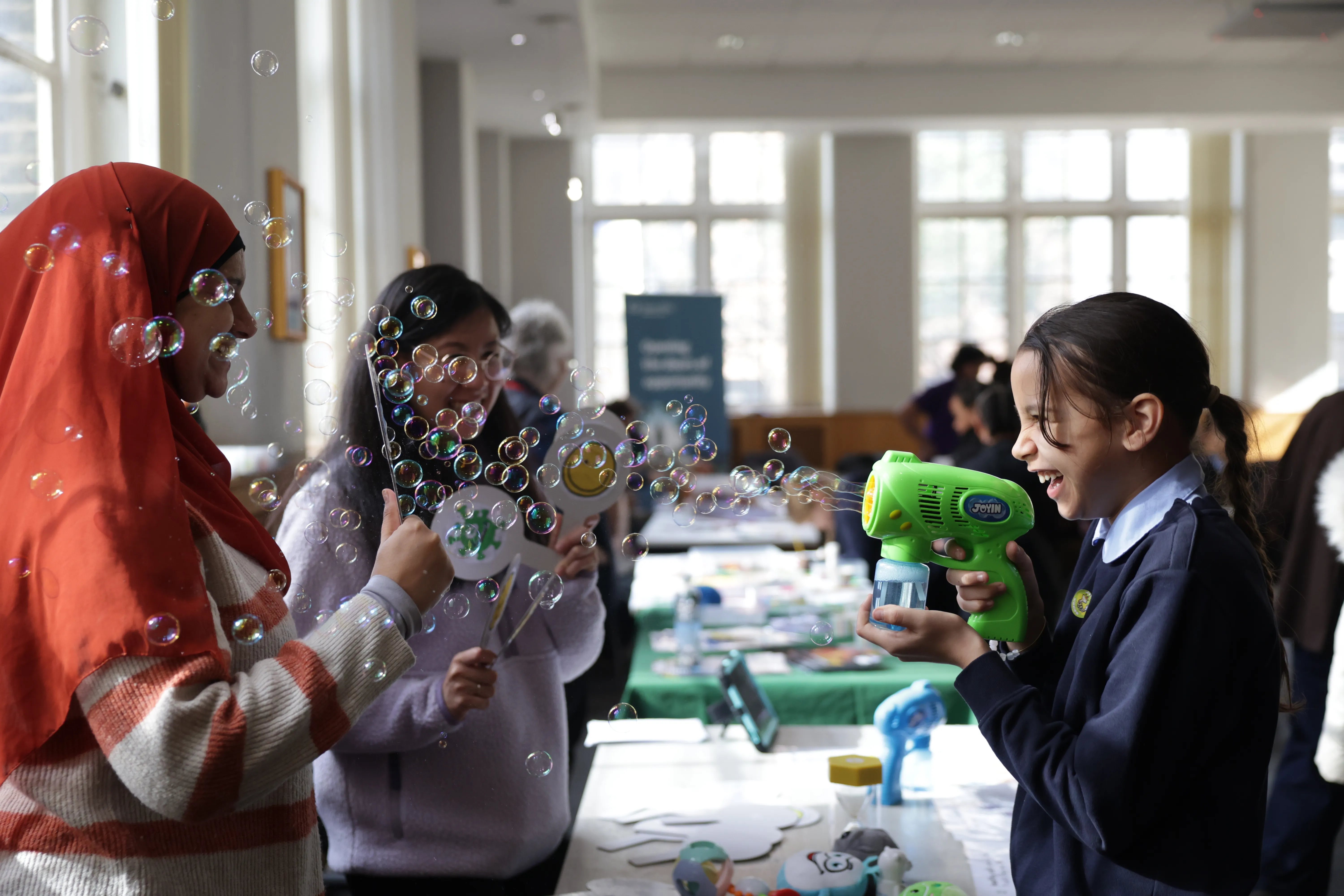 A girl blows a bubble gun at two people - they are smiling and laughing. As part of the Moon Palace STEAM activity fair at QMUL in March 2026.