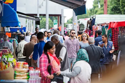 A busy street in east London: in the foreground two women chat by a market stall whilst people walk past in the background.