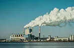 A power station next to a body of water. Smoke is billowing out of the chimneys into a blue sky.