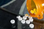 An open orange pill bottle tipped over on to a table with white tablets spilling out.