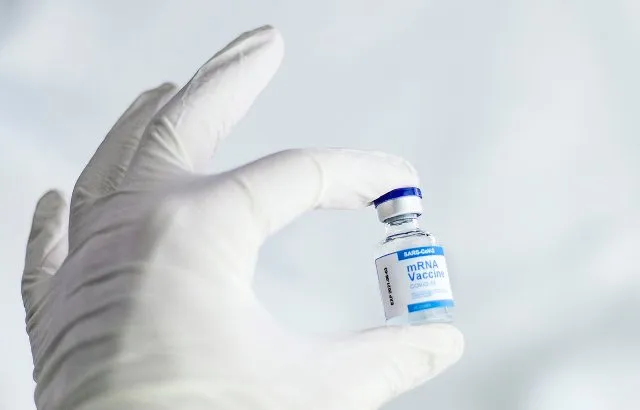 A hand wearing a white medical glove holding a small vial of mRNA Covid-19 vaccine