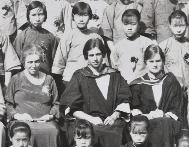 Closeup of a formal group photograph of a school. Three white women are seated centrally, the left two are young women in academic robes and the right an older woman in a dress. Standing behind and sitting in front of the women are Chinese school aged girls in a uniform all wearing corsages.