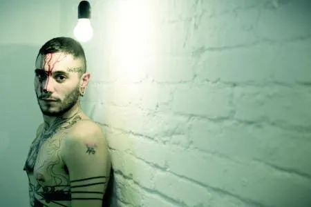 Image of Jon John standing against a white-painted brick wall. He is bare-chested and looking into the camera