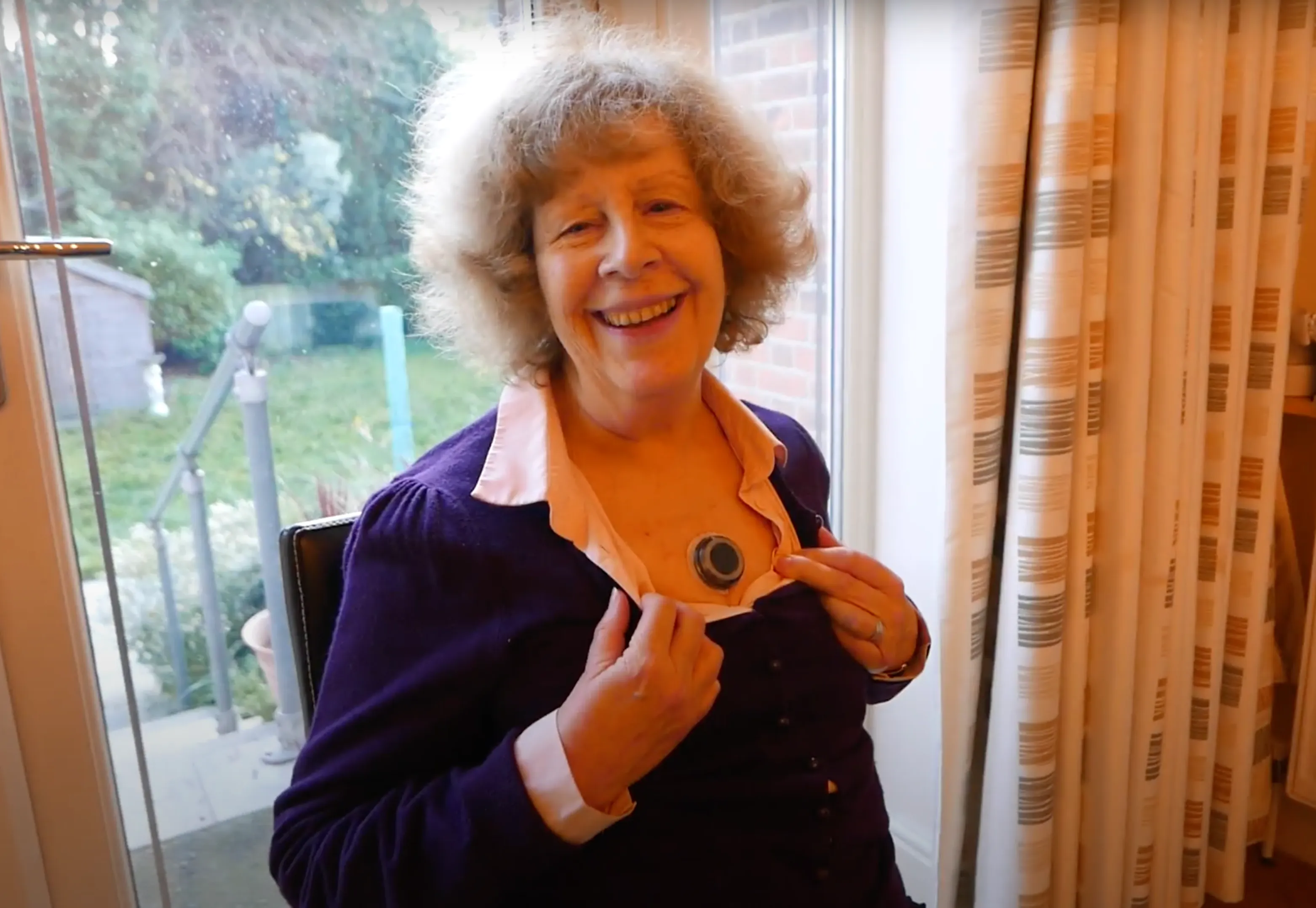 A smiling older woman pulls open the top of her shirt to show the small, black CUE1 device on her sternum.