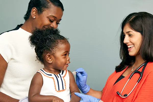 A toddler sits on her mum's knee, smiling at the doctor. The doctor is wearing gloves and is cleaning the toddler's arm ready for an injection.