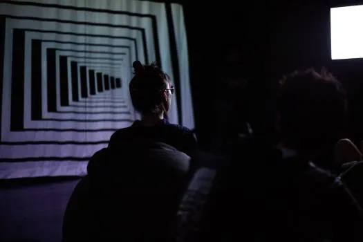 A woman sat on a beanbag in a dark space with a projecton of a geometric tunnel on the wall behind her