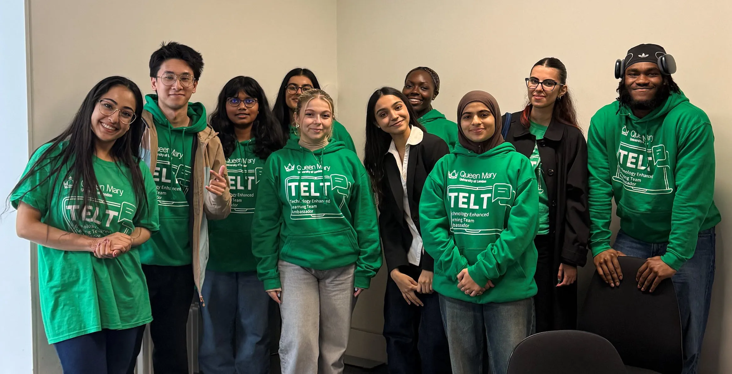 A group of TEL student ambassadors standing in front of a white wall in the corner of the room. Smiling and  looking at the camera. All wear green TELT t-shirts apart from one in a suit.