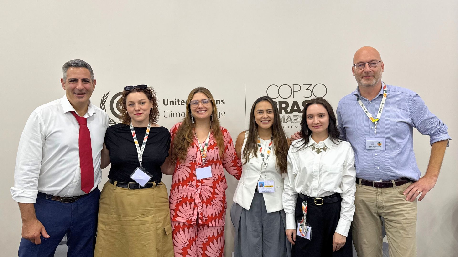 Two men and four women, all smartly dressed and smiling, standing in front of a COP30 sign on a wall in Belem in Brazil.